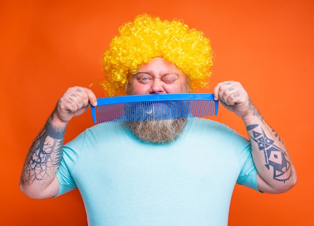 Foto grasso uomo dubbioso con barba tatuaggi e occhiali da sole si pettina con un pettine gigante