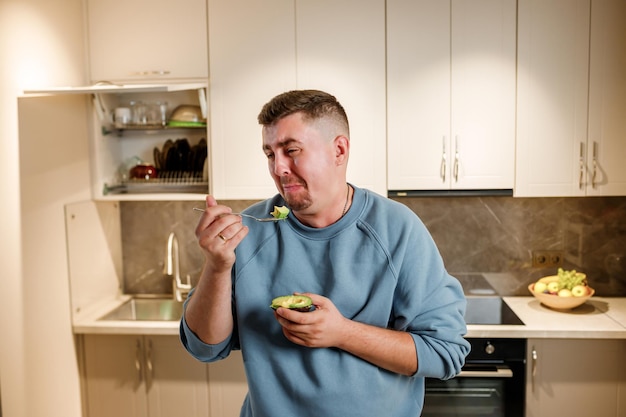 뚱뚱하고 재미있는 남자가 현대 가정 주방에 서서 슬리밍과 건강한 생활 방식 개념에 서서 아보카도를 먹습니다.