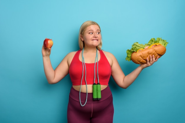 Fat girl is undecided to eat an apple or a big sandwich. cyan background