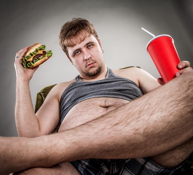 Fat man eating hamburger seated on armchair. Style fast food.