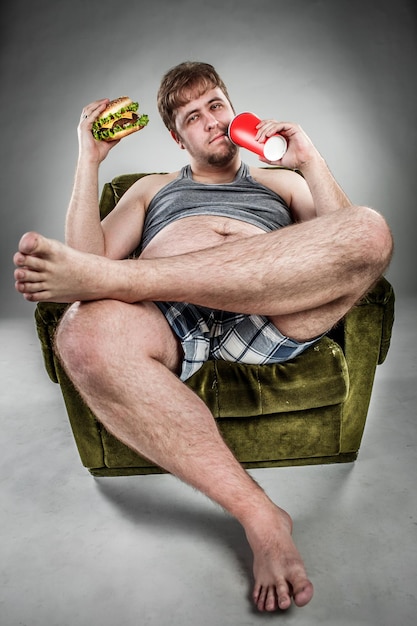 Fat man eating hamburger seated on armchair. Style fast food.