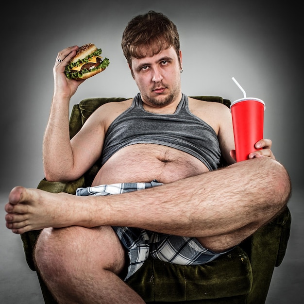 Fat man eating hamburger seated on armchair. Style fast food.