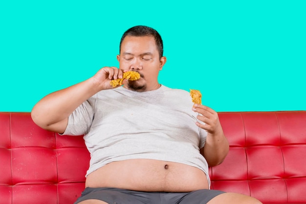 Fat man eats fast food while sitting on sofa isolated on green background