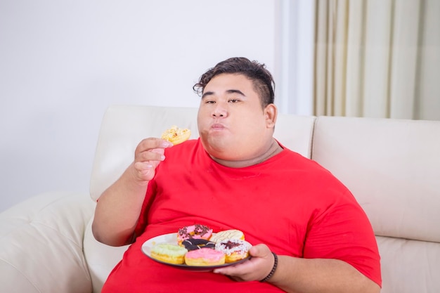 Толстый мужчина наслаждается тарелкой пончиков на диване.