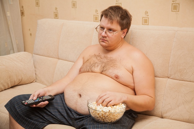 Fat man with beer belly in front of the tv eating popcorn