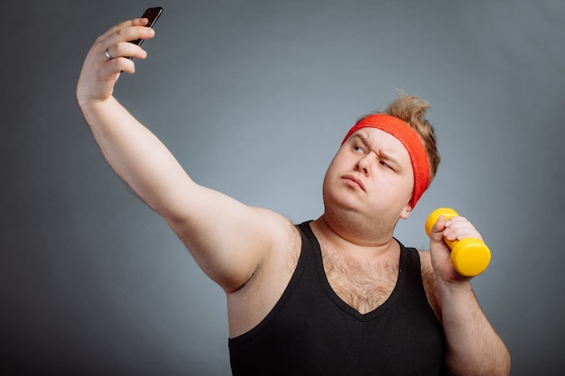 Uomo grasso con la grande pancia, tenendo il manubrio, facendo selfie sulla parete grigia