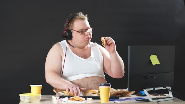 Fat man with glasses and headphones sitting at a table eating and playing a PC game at home. Self-isolation, quarantine