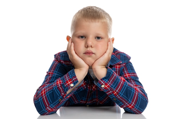 A fat, sad boy sits at a table with his head in his hands. Schoolboy in a plaid shirt. Back to school. Isolated. Close-up.