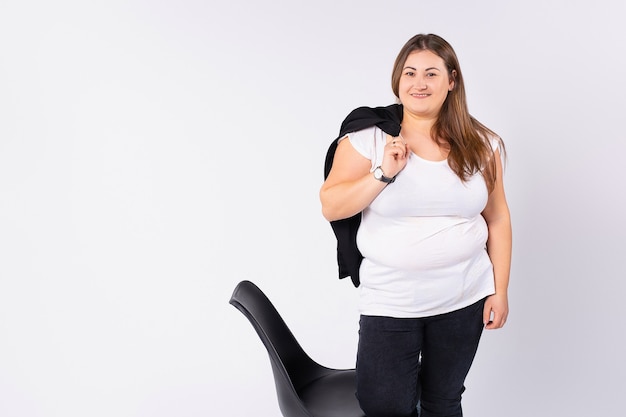 A fat woman is standing near a chair and holding a jacket in a right hand she is smiling having a good mood grey background