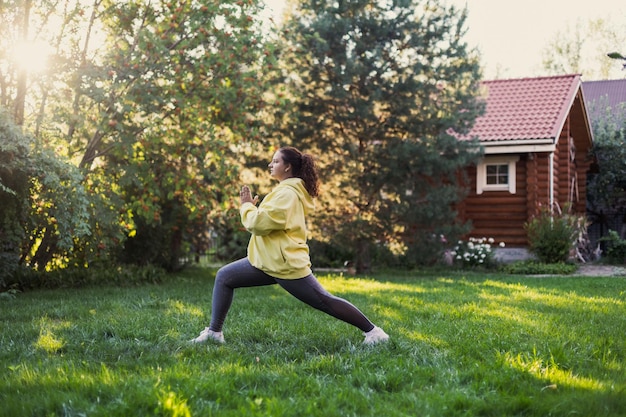 Donna grassa di mezza età che fa esercizi di yoga in abbigliamento sportivo distogliendo lo sguardo sull'erba fresca sul cortile con casa di campagna in legno e alberi ad alto fusto sullo sfondo corpo positivo