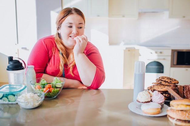 Fat young woman in kitchen sitting and eating food. Looking at sweet junk meal on left side. Temptation. Healthy food on left side.