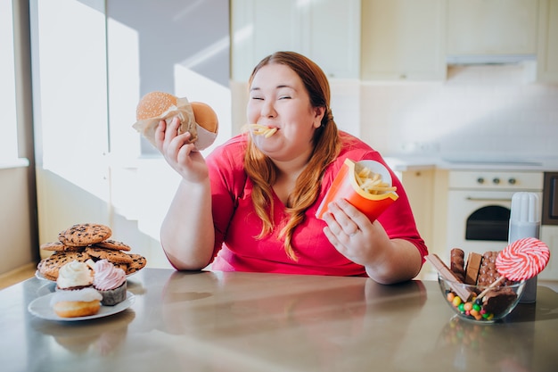 Fat young woman in kitchen sitting and eating junk food