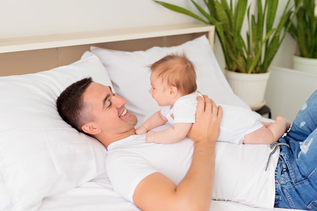 Father and baby son are playing on a white bed in a sunny bedroom.