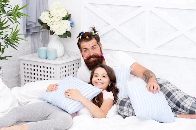 Father bearded man with funny hairstyle ponytails and daughter in pajamas. Dad and girl relaxing in bedroom. Pajamas style. Having fun pajamas party. Slumber party. Happy fatherhood. Close friends.