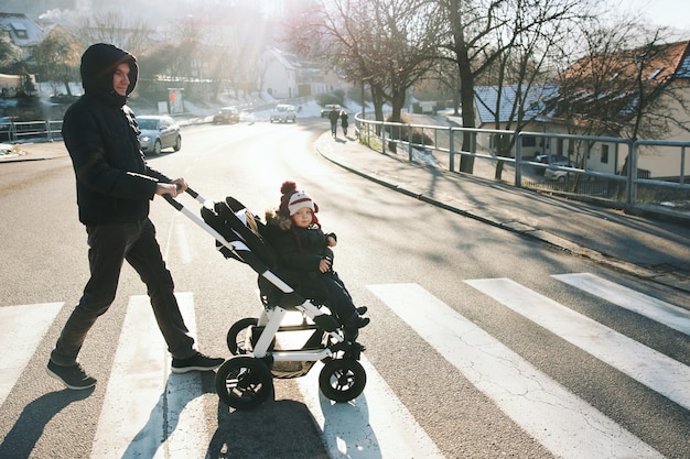 ベビーカーで変な帽子をかぶった父と少年が道を渡るヨーロッパで冬休みを過ごす家族