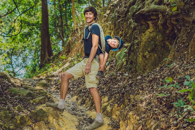 The father carries his son in a baby carrying is hiking in the forest.