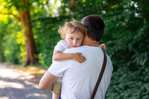 Father carries his tired little son  after a long walk in a park in summer. Sleepy exhaused toddler boy resting in daddy's arms.