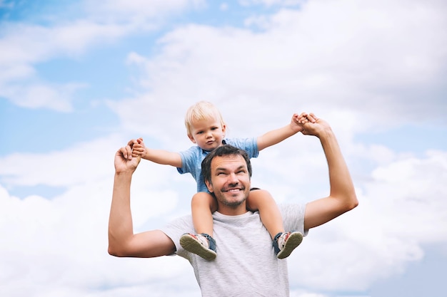 Father carrying his son on his back with raised arms up against the sky Man and child boy Family background Dad and his kid having fun at summer day on nature