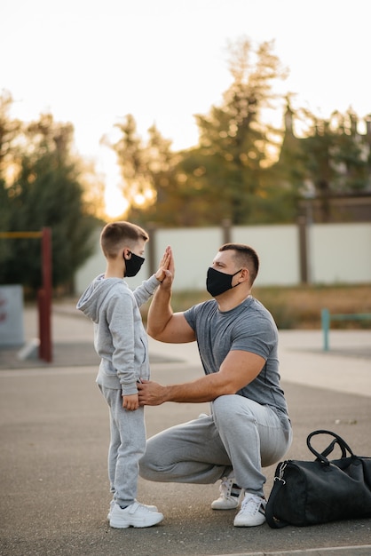 A father and child stand on a sports field in masks after training during sunset.