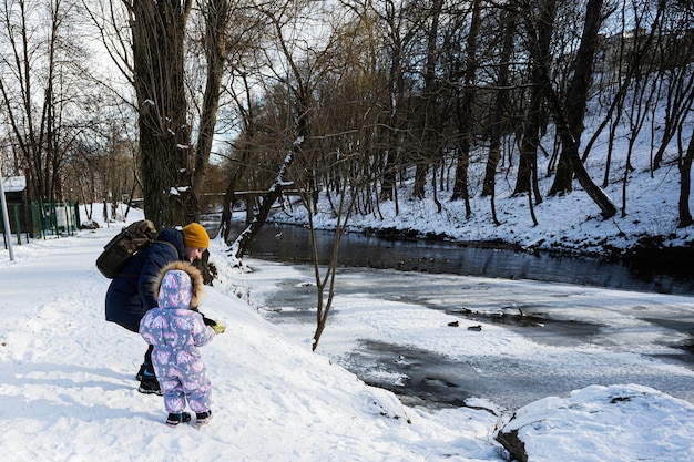 Отец и ребенок в солнечный морозный зимний день в парке кормят уток на замерзшей реке