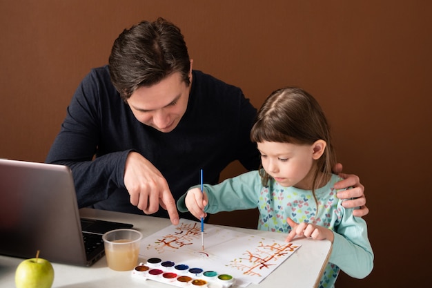 Отец и дочь рисуют дома рядом с ноутбуком