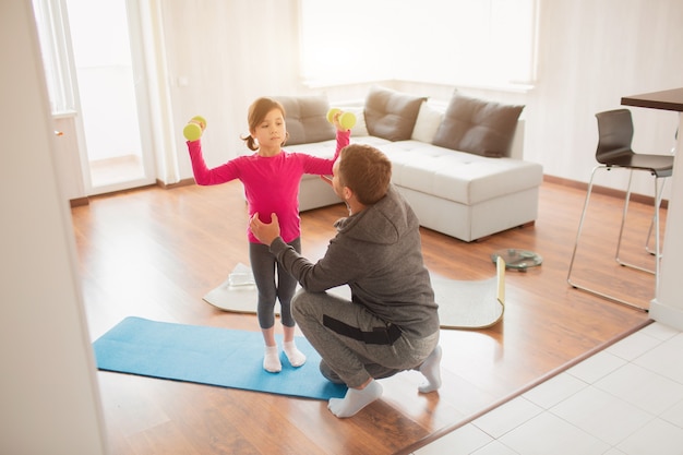 Photo father and daughter are training at home. workout in the apartment. sports at home. they are standing on a yoga mat. girl holding dumbbell.