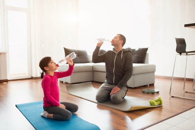 Father and daughter are training at home. Workout in the apartment. Sports at home. They sit on yoga mats and drink water. You need clean water to drink your sport time.