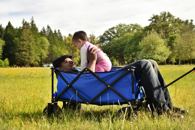Father and daughter in cart on grass against trees and sky
