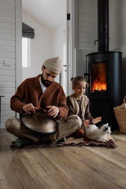 father and daughter spend time together sitting next to each other and playing handpan share home time playing with a cat and warming themselves by a hot fireplace