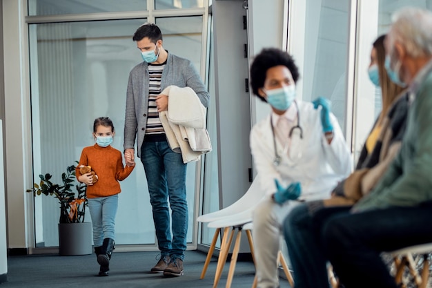 Father and daughter wearing protective face masks while holding hands and walking through hospital hallway