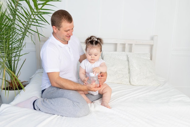 A father feeds his little daughter from a bottle on a white bed at home the concept of baby food and child care