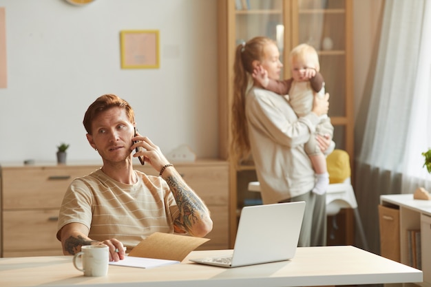 Father has a conversation on mobile phone while sitting at the table with his wife and baby  in the room