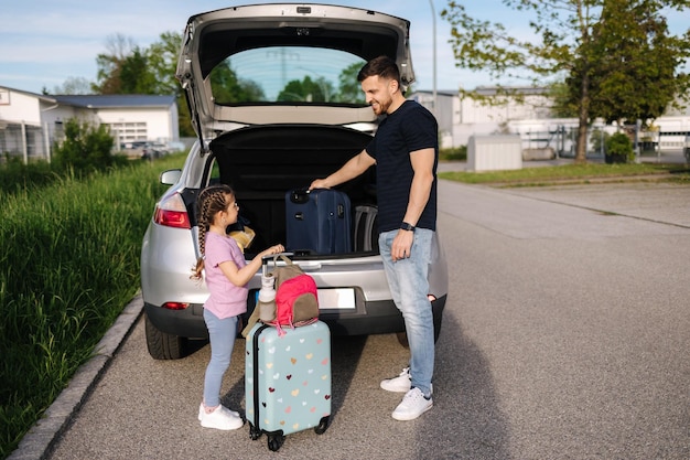 父親が旅行前に娘が荷物をトランクに積み込むのを手伝い、母親は荷物をまとめている