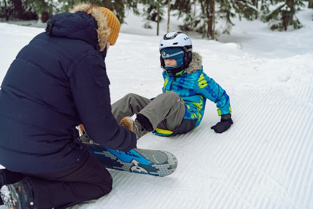 雪の上に座っている小さな男の子がスノーボードのビンディングに足を入れてストラップを調整するのを助ける父