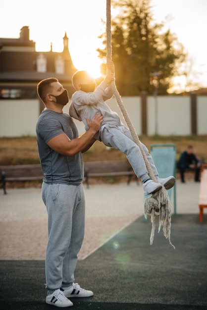 A father helps his son climb a rope on a sports field in masks during sunset