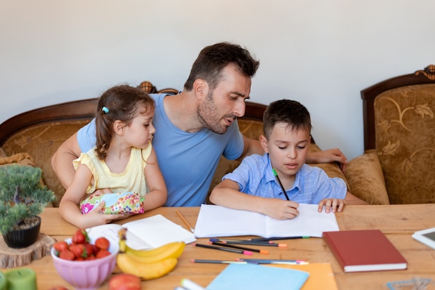 The father helps his son with school homework, makes sure that the boy writes correctly, while the youngest daughter sits in his arms.