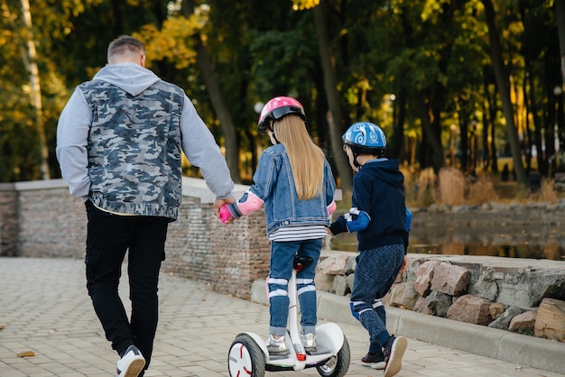 A father helps and teaches his young children to ride a Segway in the Park during sunset. Family vacation in the Park.