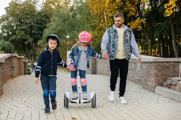 Отец помогает и учит своих маленьких детей кататься на сегвее в парке на закате.