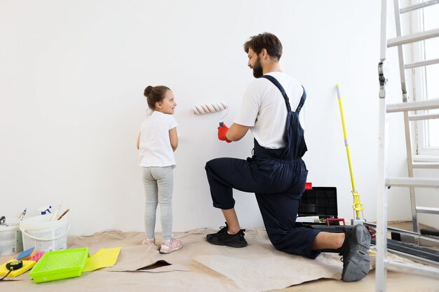 Father and his cute child daughter standing and paints the wall with white paint