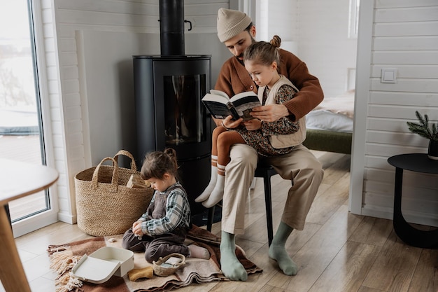 A father and his daughter are reading a book together while the other daughter is sitting on the floor at his feet and playing with toys