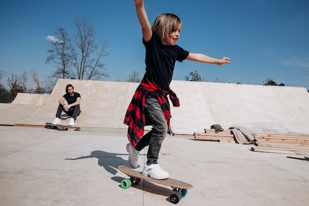 Father and his little son dressed in the casual clothes ride skateboards and have fun in a skate park with slides outside at the sunny day .