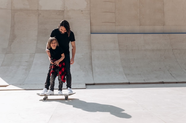 Father and his little son dressed in the casual clothes stand together on the one skateboard in a skate park at the sunny day .