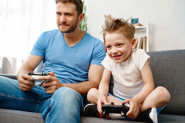 Father and his little son playing video games together on couch at home