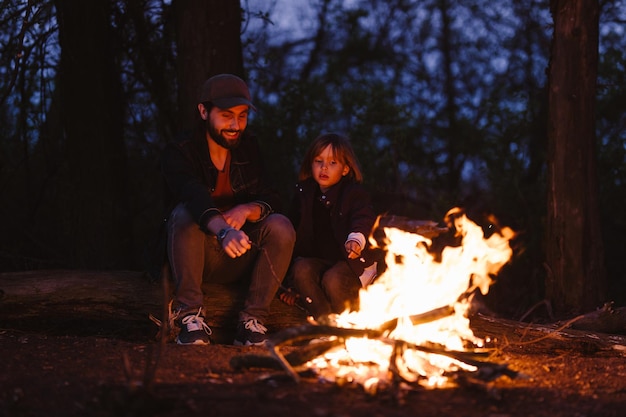 The father and his son sitting on the logs in the forest  and roasting marshmallows on the fire