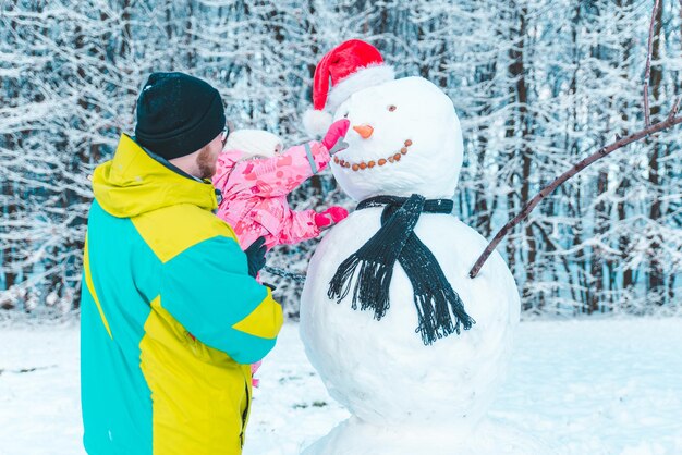 彼女が雪だるまの概念にニンジンの鼻を置くのを手伝って娘を手に持っている父