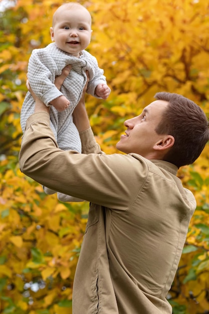 Father holds laughing baby high up against yellow trees