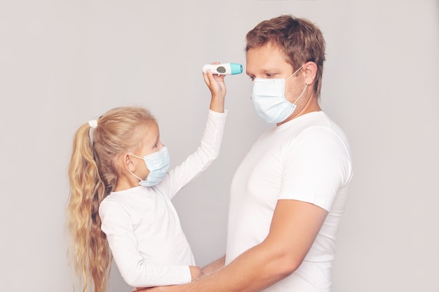 医療用マスクを着用した父親は、隔離された背景にある電子体温計で娘の体温を測定します