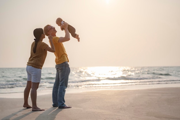 父は、休日の日没時にビーチで楽しんでいる幼児の赤ちゃんの息子と母を運んで遊んでいます。家族、ビーチ、リラックス、息子、赤ちゃん、ライフスタイルのコンセプト。