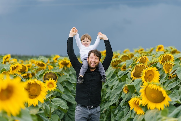 A father is having fun with his little son walking through a field with sunflowers on a summer day