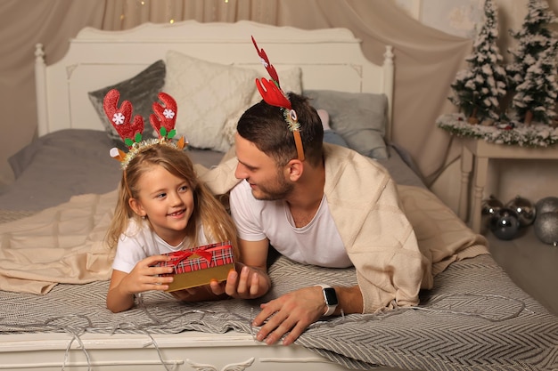 отец лежит на кровати с маленькой дочкой в ободке из оленьих рогов и смотрит на рождественскую подарочную коробку